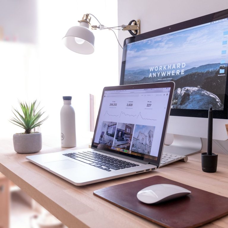 A modern desk setup with a laptop, desktop monitor, plants, and stationery items.
