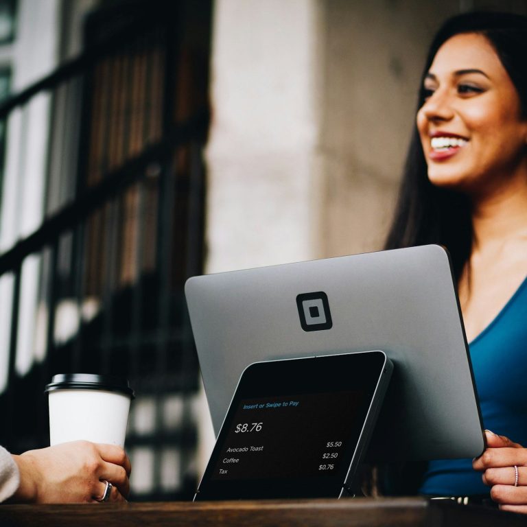 A woman smiles while working on a laptop at an outdoor cafe, holding a coffee cup.