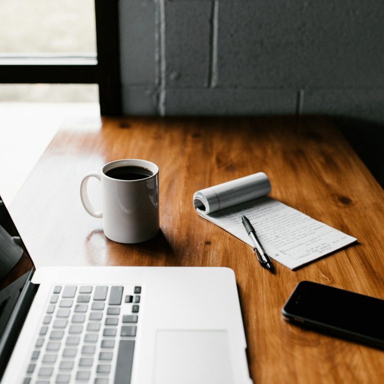 A laptop, coffee mug, notepad, and phone on a wooden table near a window.