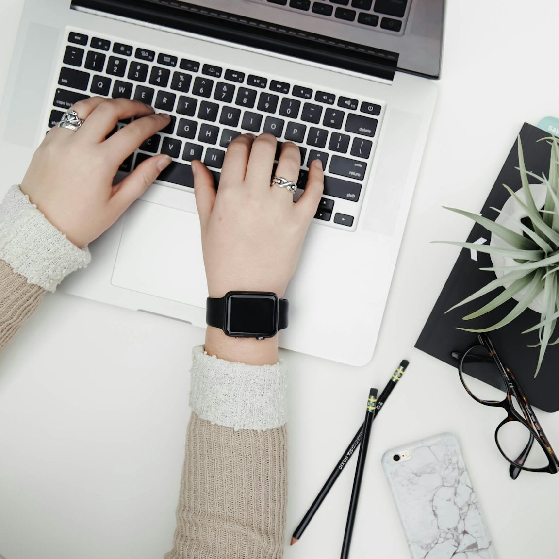Person's hands on a laptop, with a plant, glasses, phone, and pens nearby.